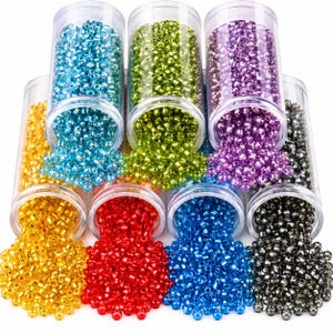Puede incluir: Un conjunto de ocho recipientes cilíndricos de plástico transparente llenos de pequeñas cuentas de semillas con acabado metálico en varios colores, incluyendo amarillo, azul claro, verde lima, morado, rojo, azul oscuro y negro, derramándose.