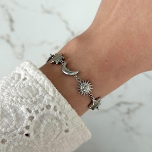 May include: A silver-toned metal chain bracelet featuring alternating charms of stars, a crescent moon, and a sun with a face. The bracelet is worn on a wrist, showcasing the polished metallic finish and the detailed celestial-themed charms.