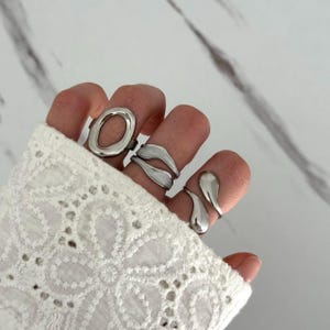 May include: A set of three silver-toned rings worn on fingers. One ring features a large oval shape, another is a double-banded design with a smooth finish, and the third is a teardrop-shaped open ring. The jewelry has a polished metallic surface.