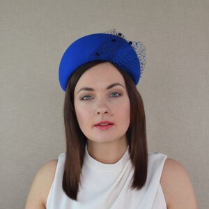 May include: A bright blue felt hat with a black netting detail. The hat is shaped like a half-circle and sits on top of the head.