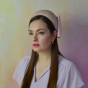 Op de afbeelding: Een vrouw draagt een lichtroze hoofdband met een strik aan de zijkant. De hoofdband is gemaakt van een zacht, fluweelachtig materiaal. De vrouw heeft lang bruin haar en draagt een lichtroze blouse.