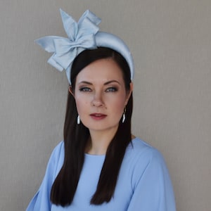 May include: A light blue headband with a large bow on top. The headband is made of satin and has a padded band. The model is wearing the headband on her head and has long dark hair.