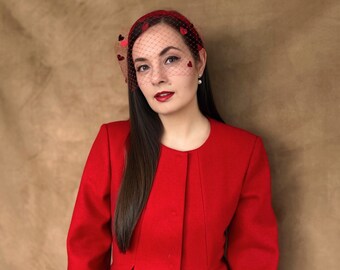 May include: A woman wearing a red jacket with a red veil headband with heart-shaped cutouts. The veil is covering her forehead and hair.