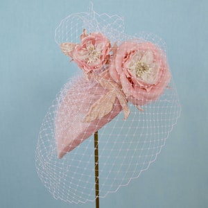 May include: A pink hat with a large pink flower and a smaller pink flower on top. The hat is decorated with gold glitter and is covered in a sheer pink veil.