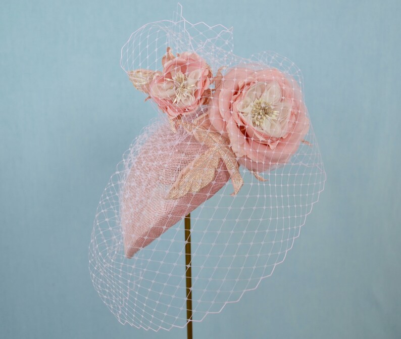 May include: A pink hat with a large pink flower and a smaller pink flower on top. The hat is decorated with gold glitter and is covered in a sheer pink veil.