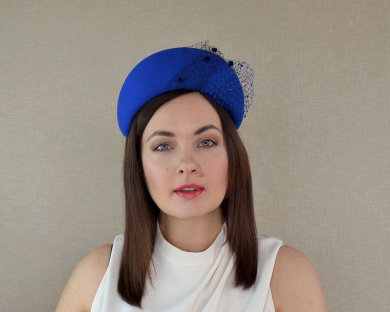 May include: A royal blue felt hat with a black netting detail. The hat is shaped like a half-circle and sits on the head.