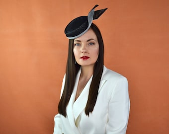 Op de afbeelding: Een vrouw draagt een wit blazer met een zwarte en zilveren hoed. De hoed heeft een kleine rand en een lang, golvend lint.