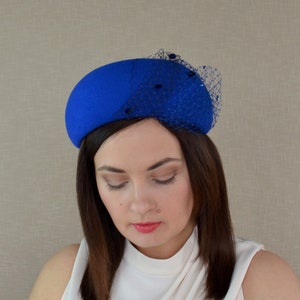 May include: A royal blue felt hat with a black netting and small black beads. The hat is tilted slightly to the side.