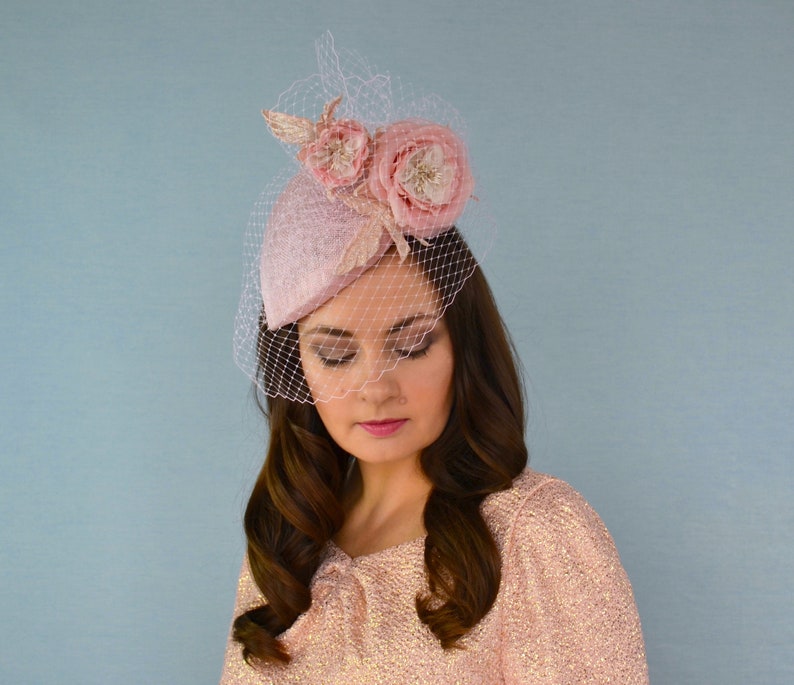 May include: A pink hat with a veil and two pink fabric flowers. The hat is tilted slightly to the side.