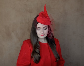 May include: A woman wearing a red dress and a red hat with a veil. The hat has a feather detail.