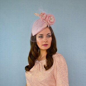 May include: A pink felt hat with a veil and two pink fabric flowers. The hat is tilted on the head of a person with long brown hair.