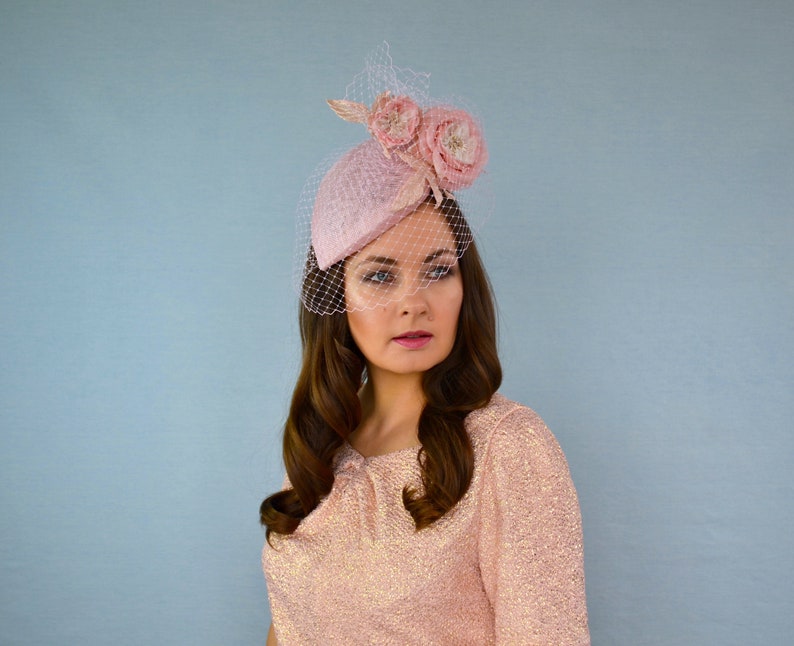May include: A pink felt hat with a veil and two pink fabric flowers. The hat is tilted on the head of a person with long brown hair.