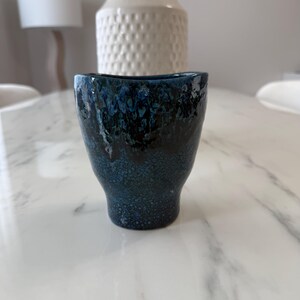 Peut inclure: Une petite tasse en céramique avec un émail réactif bleu profond et noir à la texture mouchetée. Le récipient présente un bord légèrement évasé et une base effilée, avec une finition brillante passant de l'indigo foncé au bleu clair.
