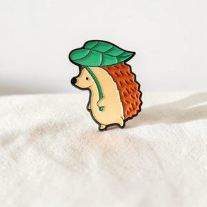 May include: A metal enamel pin shaped like a small hedgehog with a cream-colored face and belly, brown textured quills, and a large green leaf held over its head like an umbrella. The design features bold black outlines throughout the entire figure.