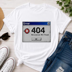 May include: A white short-sleeved t-shirt featuring a graphic print of a computer error message box. The box displays the text Error, a red circle with a white X, the number 404, the text Motivation Not Found, and an OK button.