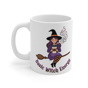 May include: A white ceramic mug featuring a cartoon illustration of a witch with purple hair wearing a black hat, sitting on a broomstick while holding a coffee cup with a pumpkin design. The text Basic Witch Energy is printed below.