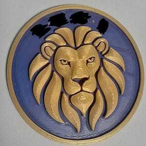 May include: A circular wall plaque featuring a three-dimensional gold-colored lion head relief centered on a flat, textured purple background. The lion has a detailed mane and facial features. The plaque is framed by a raised gold-colored circular border.