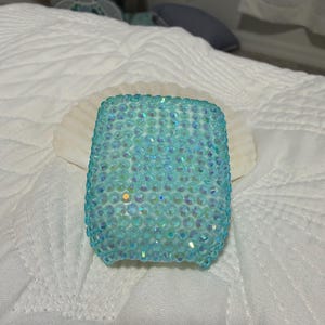 Può includere: Una custodia protettiva rettangolare per auricolari wireless, interamente ricoperta di piccoli strass azzurri iridescenti che creano una texture superficiale scintillante e sfaccettata sull'intera parte anteriore e sui lati della custodia.