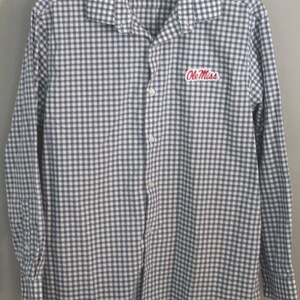 May include: A long-sleeve button-down shirt featuring a navy blue and white gingham check pattern. The shirt has a standard collar, a front button placket, and the text Ole Miss embroidered in red script on the upper left chest area.