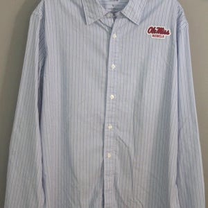 May include: A long-sleeve button-down shirt featuring a light blue and white vertical pinstripe pattern. The shirt has a classic collar and a red and white embroidered patch on the left chest that reads Ole Miss REBELS in a script font.