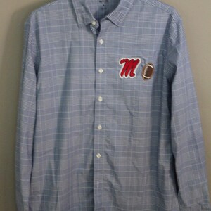 May include: A light blue and white plaid long-sleeve button-down shirt featuring a red script letter M patch and a brown football patch on the left chest. The shirt has a standard collar, a full front button placket, and long sleeves with cuffs.
