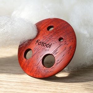 Puede incluir: Un botón circular de madera de color marrón rojizo con un patrón de veta de madera distintivo y cuatro agujeros de diferentes tamaños. La palabra "Fotool" está grabada en letras negras en la superficie del botón, parcialmente cubierto por fibra blanca.
