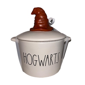 May include: A cream-colored ceramic canister with a lid featuring a brown sculpted sorting hat topper. The canister has two side handles and the word HOGWARTS printed in black, sans-serif lettering on the front. A white ribbon is attached to the lid.