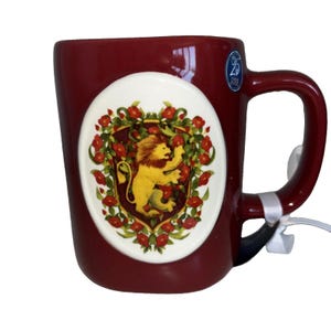 May include: A deep maroon ceramic mug featuring a central oval emblem with a yellow lion illustration surrounded by red flowers and green leaves. A small blue circular sticker with the text 25 Years of Magic is visible near the top right handle.