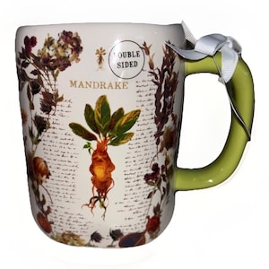 May include: A cream-colored ceramic mug with a green handle and a white ribbon tied to it. The mug features a botanical illustration of a mandrake root, various dried flowers, and cursive script text. A circular label reads DOUBLE SIDED.