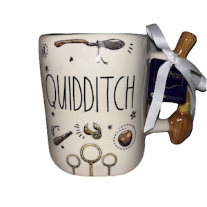 May include: A cream-colored ceramic mug featuring the word QUIDDITCH in black lettering, surrounded by illustrations of a broomstick, a golden snitch, a telescope, and three hoops. The handle is shaped like a broomstick with a white ribbon attached.