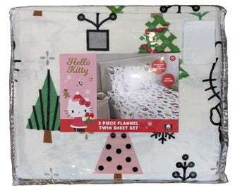 May include: A 3-piece flannel twin sheet set in white featuring a Hello Kitty holiday print with green and pink Christmas trees, snowflakes, and gifts. The packaging includes a product image showing the sheets on a bed and the text Hello Kitty.