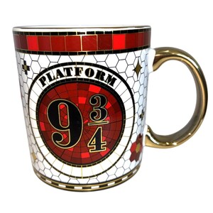 May include: A white ceramic mug with a gold-colored handle featuring a circular design that reads PLATFORM 9 3/4 in black and gold lettering. The mug has a red and black mosaic-style border and a white honeycomb pattern background with gold stars.