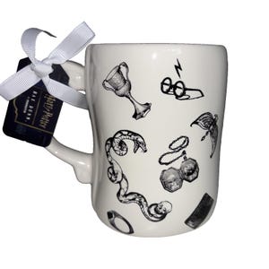 May include: A white ceramic mug featuring black ink illustrations of a goblet, round glasses with a lightning bolt, a snake, a locket, a winged key, a ring, and a skull. A white ribbon is tied to the handle with a Harry Potter Rae Dunn tag.