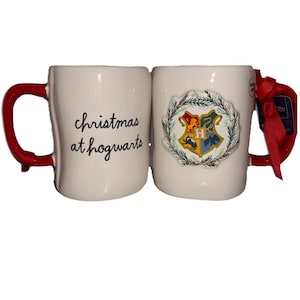 May include: Two white ceramic mugs with red handles. One mug features the black text "christmas at hogwarts" in a cursive font. The other mug displays a colorful Hogwarts crest emblem surrounded by a green and red holiday wreath design.