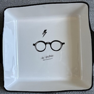 May include: A square white ceramic baking dish with black trim and handles. The center features a black graphic of round glasses and a lightning bolt above them, with the text Harry Potter printed in black below the glasses on the white surface.