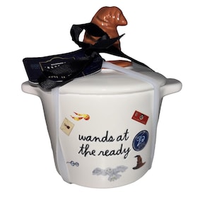 May include: A white ceramic lidded jar with a brown hat-shaped handle. The jar features the text wands at the ready and small illustrations of a golden snitch, a letter, glasses, an owl, a hat, and a red ticket. A black ribbon and tag are attached.