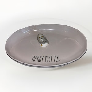 May include: A light gray oval ceramic trinket dish featuring a small, sculpted gray owl figurine attached to the center. The text HARRY POTTER is printed in black, handwritten-style lettering on the surface of the dish below the owl figure.