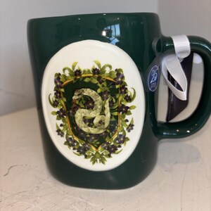 May include: A dark green ceramic mug featuring a central oval emblem with a white background. The emblem displays a coiled light-colored snake surrounded by green leaves and purple flowers. A white ribbon is tied to the handle with a small tag.