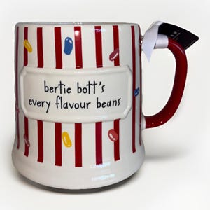May include: A white ceramic mug featuring vertical red stripes and colorful jelly bean illustrations. A central raised rectangular plaque displays the black text "bertie bott's every flavour beans". The mug has a solid red handle with a white ribbon tag.