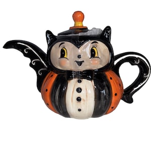 May include: A ceramic teapot shaped like a character with a black and orange color scheme. The teapot features a black bow tie, a white front panel with three black dots, a curved handle, and a spout shaped like a wing. The lid has an orange knob.