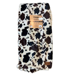 May include: A Wrangler brand faux fur micro mink throw blanket featuring a black and white cow print pattern. The blanket includes a rectangular product information tag and a small brown leather-like patch with the Wrangler logo at the bottom.