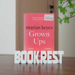 Puede incluir: Un libro rosa titulado Grown Ups de Marian Keyes colocado detrás de un soporte blanco con letras de bloque que forman la palabra BOOK REST. La portada del libro presenta texto en negro y una pequeña etiqueta blanca con un código de barras.