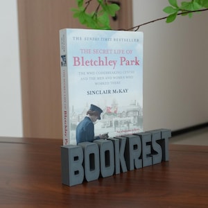 Puede incluir: Un sujetalibros de hormigón gris con la palabra BOOKREST en letras de bloque, sosteniendo un ejemplar de The Secret Life of Bletchley Park de Sinclair McKay. La portada del libro muestra una ilustración histórica de una persona en uniforme.