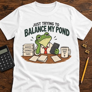 May include: A white t-shirt featuring a green frog wearing a white shirt and red tie, sitting at a desk with a calculator, a stack of papers labeled INVOICES, EXPENSES, and TAXES, and a mug labeled COFFEE ADDS UP. Text reads JUST TRYING TO BALANCE MY POND.