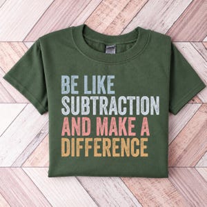 May include: A dark olive green short-sleeve t-shirt featuring the distressed-style text "BE LIKE SUBTRACTION AND MAKE A DIFFERENCE" printed in light blue, light pink, and yellow-orange block lettering across the center of the chest.