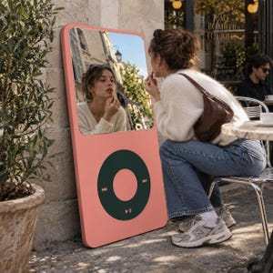 May include: A tall, rectangular floor mirror designed to resemble a vintage digital music player. The frame is coral pink with a large, dark green circular control interface featuring playback icons at the bottom and a reflective glass mirror surface.