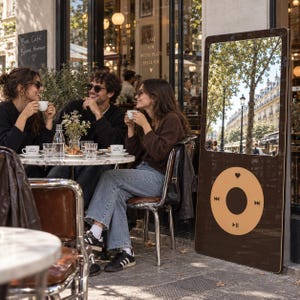 May include: A tall, rectangular, dark brown mirror designed to resemble a vintage digital music player. The mirror features a large reflective upper section and a lower control panel with a circular interface, including a heart icon, play-pause symbol, and track skip icons in a light tan color.