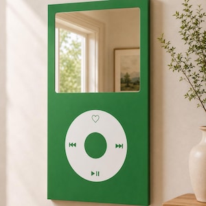 May include: A rectangular wall mirror with a green frame designed to resemble a digital music player. The upper portion is a reflective glass surface, while the lower section features a white circular control interface with icons for play, pause, skip, and a heart symbol.