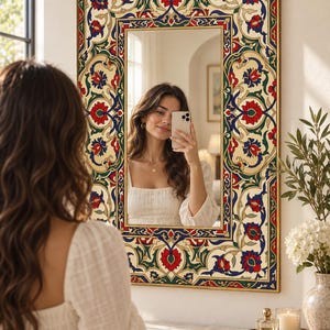 May include: A rectangular wall mirror featuring a wide, ornate frame decorated with a colorful floral pattern in shades of red, blue, and green against a cream background. The mirror has a thin gold-toned border surrounding the glass surface.