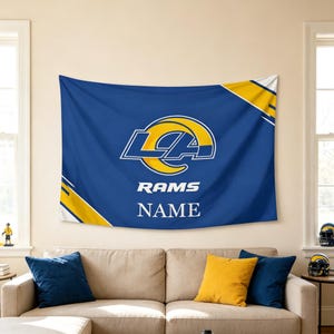 May include: A rectangular blue wall tapestry featuring the Los Angeles Rams logo in yellow and white, with the word RAMS printed below the logo and the word NAME printed at the bottom. The design includes yellow diagonal stripes in the corners.